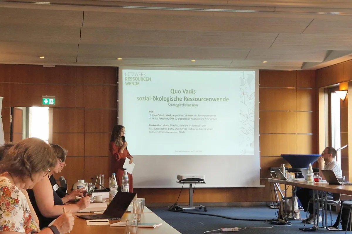In einem Konferenzraum sitzen Menschen an Tischen. Am Whiteboard steht eine Moderatorin. 